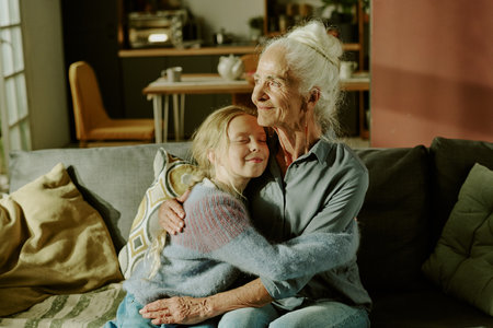 Senior woman with white hair embracing young girl on couch in cozy living room filled with natural light, creating heartwarming moment of affection and family bondの写真素材