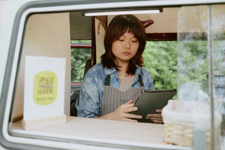Asian female food truck worker using tablet to check new orders while sitting inside of vanの写真素材