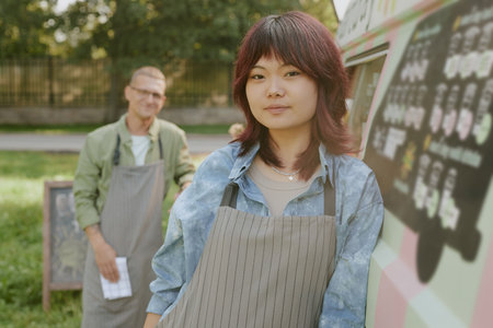 Medium close up portrait of Asian female worker looking at camera and smiling while her colleague in...