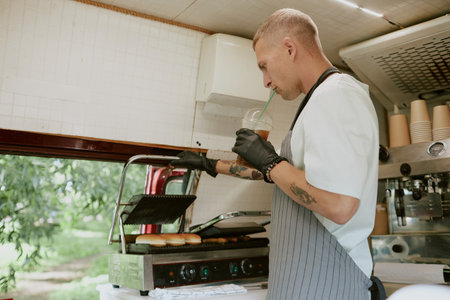 Young adult chef drinking cold brew coffee while checking buns for hot dogs on grillの写真素材