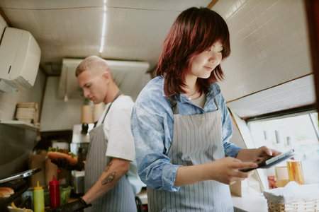 Low angle shot of Asian female cook checking new orders in app on tablet while Caucasian male chef preparing hot dogs in blurred backgroundの写真素材