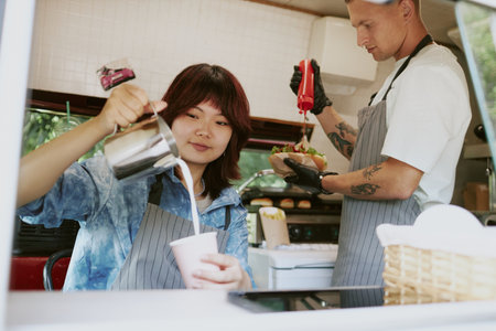 Asian female barista adding milk into fresh brewed coffee while Caucasian male chef adding ketchup in hot dog in backgroundの写真素材