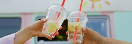 Website banner with unrecognizable couple clinking transparent plastic gups with citrus lemonade and slices of lime insideの写真素材