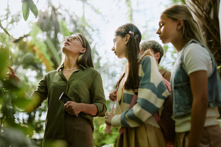 Low angle view of teen students visiting botanical garden, listening to female specialist speaking...