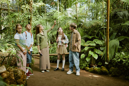 Wide shot of young female botanical garden worker demonstrating various plants growing in greenhouse to group of teen studentsの写真素材