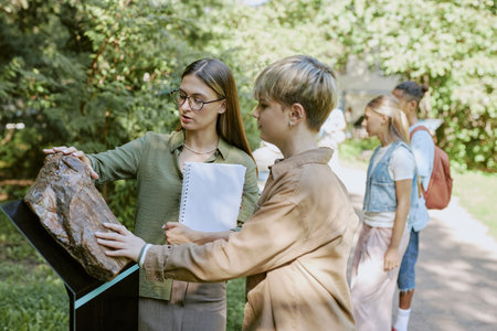 Female botanical garden worker showing petrified tree log to teen boy during tour, medium shotの写真素材