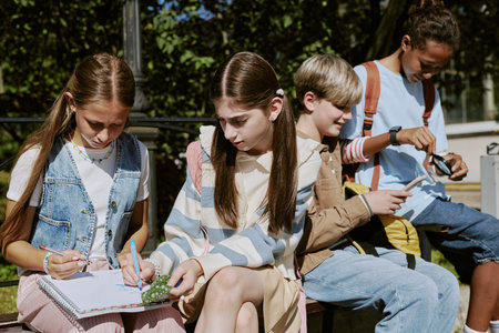 Medium shot of teen girls sitting on bench drawing in sketchbook, their classmates using smartphone and magnifier in backgroundの写真素材