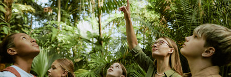 Young female botanical garden worker pointing finger at palm tree while conducting tour for group of...