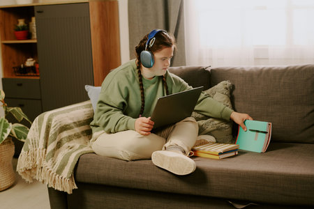 Girl with Down syndrome wearing headphones sitting on sofa with laptop on her lap, taking copybooks...