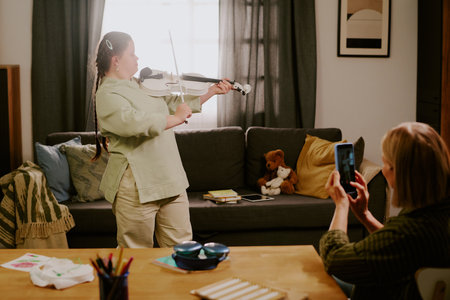 Caucasian girl with Down syndrome playing music on violin in front of her senior mother, who taking photos of her on smartphoneの写真素材