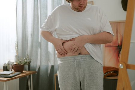 Man examining his stomach in casual home setting with natural light filtering through the curtains. Room features cozy decor with a small table and indoor plantsの写真素材