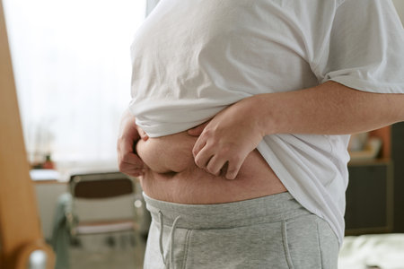 Person holding up shirt to reveal belly while standing in a room with furniture in the background,...