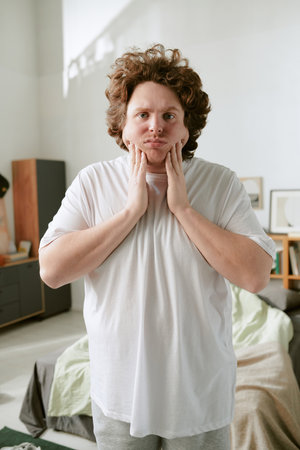 Portrait of man wearing casual attire, standing in modern apartment, holding face and appearing pensive. Furniture and home decor visible in backgroundの写真素材