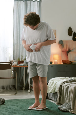 Man wearing casual home attire standing in a bedroom with modern furnishings around. He is touching his shirt, appearing relaxed in a calm home environmentの写真素材