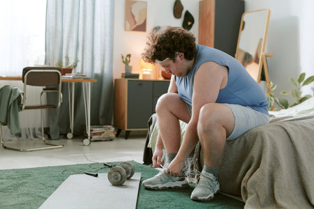 Man in casual workout attire taking a break after exercise in modern home setting. Dumbbells and resistance bands on mat in foreground with desk and plants in backgroundの写真素材