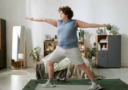 Man performing indoor exercise with outstretched arms, standing on a yoga mat in a well-decorated room near dumbbells and plantsの写真素材