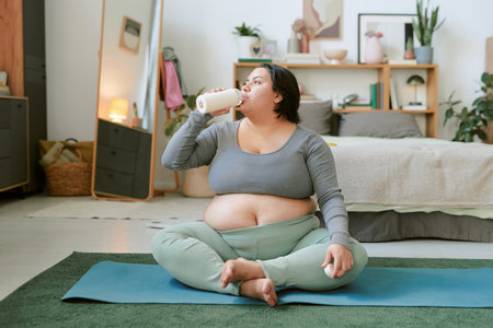 Pregnant woman sitting on yoga mat in cozy bedroom, drinking milk from bottle, surrounded by soft pastel tones and home furnishingsの写真素材