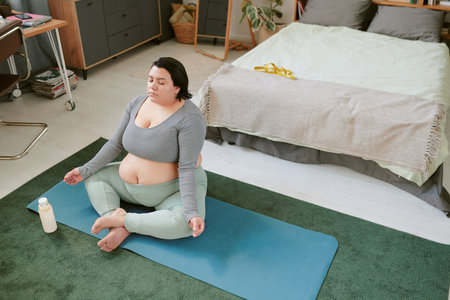Woman practicing yoga and meditation on blue mat in a cozy room with bookshelf in background and a...