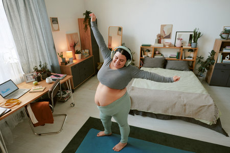 Female doing yoga exercise with headphones in cozy bedroom. Home office visible with laptop, documents, and plants creating a relaxing environmentの写真素材