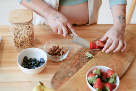 Cutting fresh strawberries and preparing a nutritious breakfast bowl with almonds, blueberries, and granola on wooden tableの写真素材