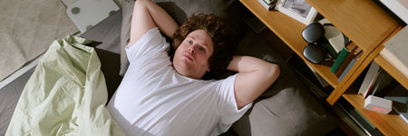 Portrait of person lying on bed with arms behind head, surrounded by books and electronics, creating a cozy and introspective atmosphereの写真素材