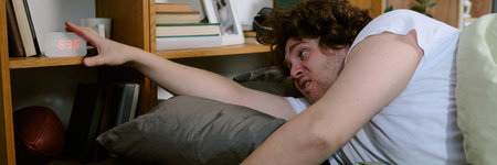 Young man with curly hair reaching to silence alarm clock in morning bedroom. Partially awake and groggy expression while lying on bed with wooden bookcase in backgroundの写真素材