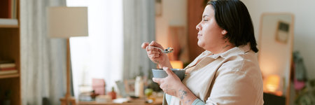 Person enjoying breakfast while standing in cozy, well-lit home environment filled with furniture and warm lighting elements, showcasing calm and content expressionの写真素材