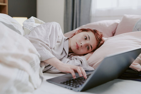 Portrait of woman lying in bed and using laptop computer, appearing relaxed and engaged. Lightly resting head on pillow, engaging with technology, and creating a serene atmosphereの写真素材