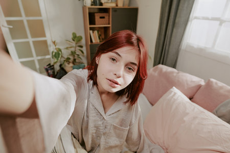 Portrait of a woman with red hair sitting on bed and wearing a relaxed expression. Background includes wardrobe, bookshelf, window with sheer curtains, and various cozy elementsの写真素材
