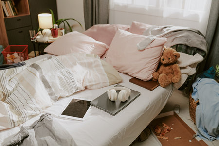 Bedroom featuring soft pillows, a teddy bear on the bed, and various personal items scattered around. Sunlight illuminating the space through a window, creating a warm atmosphereの写真素材