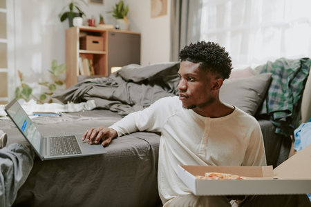 Man is using laptop while sitting on bed in cozy room filled with natural light, with soft furniture and shelves in background. Scattered objects on bed add a casual atmosphereの写真素材
