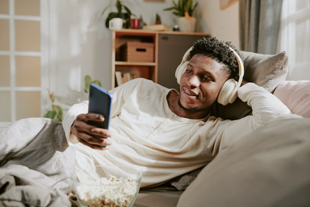 African American man lying on couch wearing headphones and holding a smartphone while leaning back. Cozy living room environment creating a comfortable and inviting atmosphereの写真素材