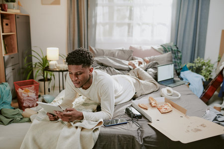 Young man relaxing on bed while using tablet in a cluttered room filled with personal items and...