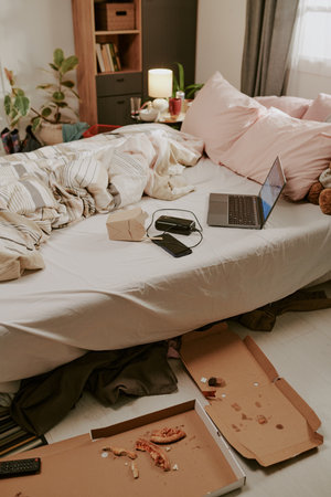 Disorganized bedroom with unmade bed, scattered pillows, open laptop, and pizza boxes. Shelves and dresser filled with books and decor items in backgroundの写真素材