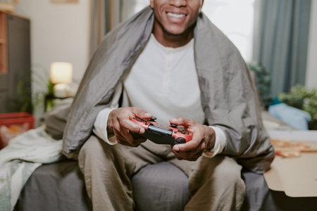 Portrait of smiling young man enjoying video gaming while seated on sofa wrapped in blanket, holding...