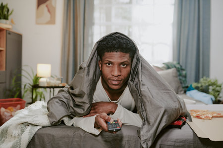 Man sitting on bed wrapped in blanket while holding TV remote, creating a relaxed atmosphere in cozy...