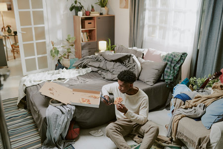 Multiracial man sitting on floor, painting in sketchbook in a comfortably cluttered living room filled with personal belongings and natural decor elementsの写真素材