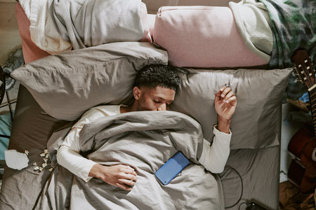 Man lying in bed holding smartphone while resting, with comfortable pillows and sheets surrounding...