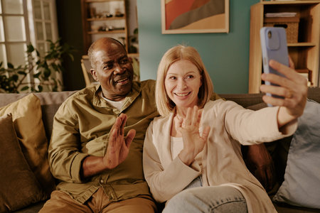 Senior Caucasian woman and senior Black man sitting on sofa smiling and waving while holding...