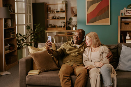 Senior Black man and senior Caucasian woman sitting on sofa taking selfie together, smiling and...