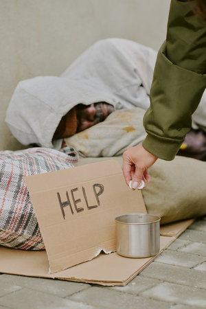 Black man sleeping on sidewalk under blanket with cardboard sign reading help while Caucasian woman...