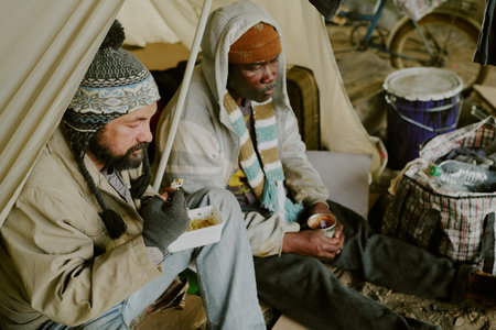 Middle aged Caucasian man and middle aged Black man sitting together under tent, eating food from...