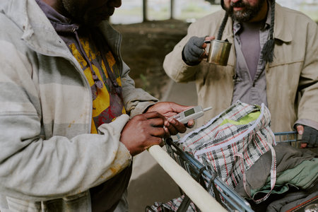Middle aged Black man holding mobile phone next to middle aged Caucasian man drinking from metal cup, both standing by shopping cart filled with personal belongings outdoorsの写真素材
