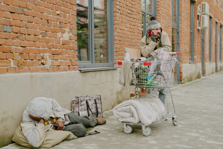 Middle aged Black man lying on sidewalk resting head on pillow near shopping bag, while middle aged Caucasian man standing beside shopping cart talking on smartphone outside brick buildingの写真素材
