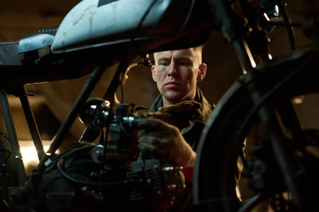 Caucasian young adult man repairing custom motorcycle in workshop, focusing on engine components...