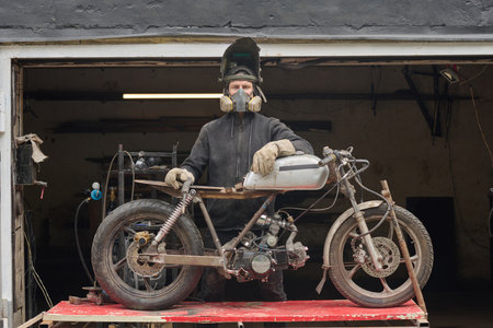 Middle aged Caucasian man wearing protective mask and gloves repairing custom motorcycle in workshop...