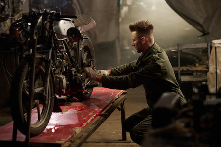 Young adult man kneeling beside custom motorcycle repairing engine components in workshop or garage,...