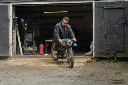 Caucasian middle aged man repairing custom motorcycle in workshop garage, sitting on bike while...