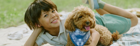 Portrait of biracial child boy lying on picnic blanket outdoors smiling at camera, with curly brown dog wearing blue bandana relaxing together in park on sunny dayの写真素材