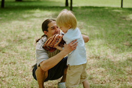 Asian young adult man crouching and hugging his child boy outdoors in park, man holding baseball...